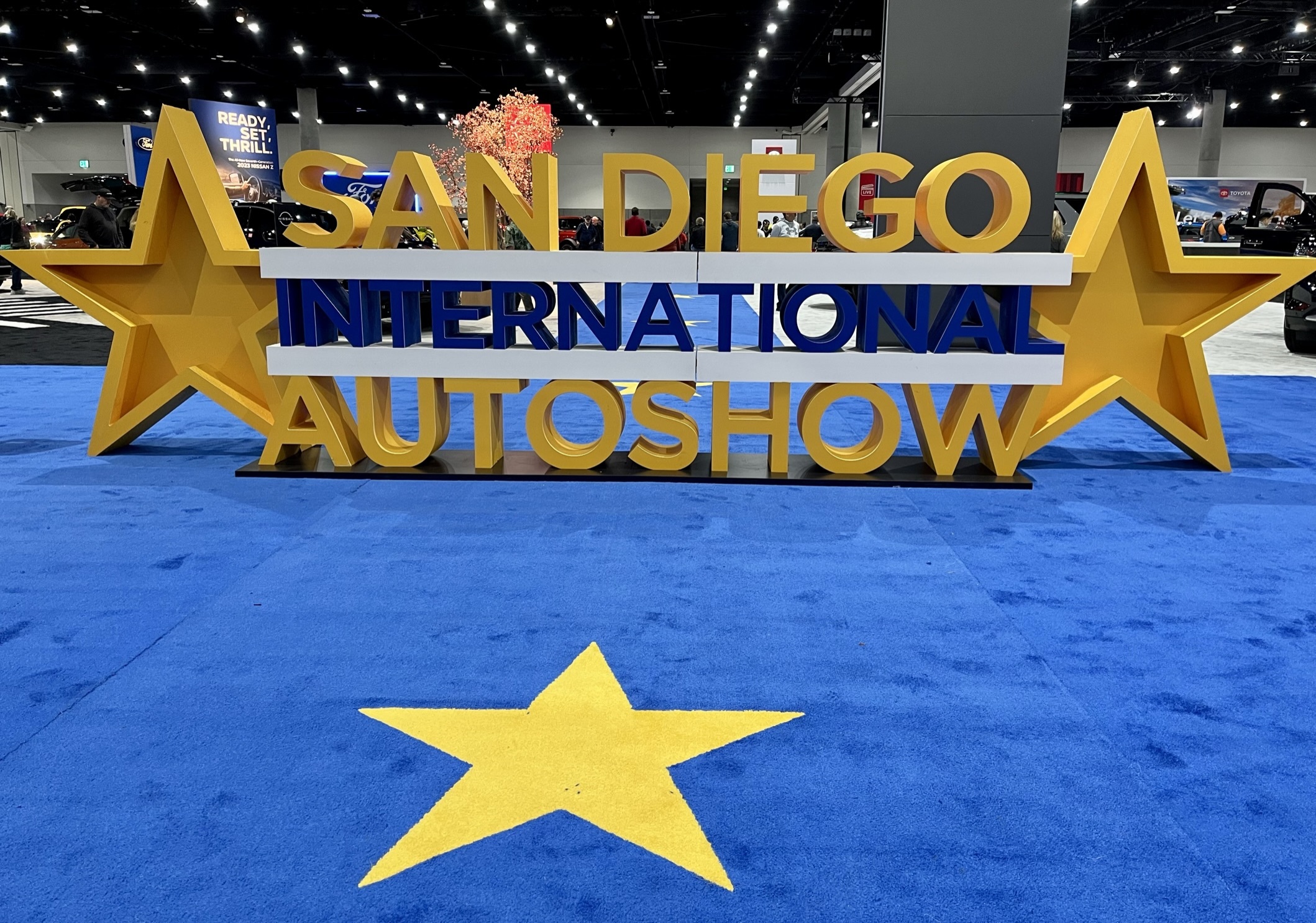 The cars are the stars at the San Diego International Auto Show. Image shows a big gold star on royal blue carpet with a stacked three-row icon for San Diego International Auto Show