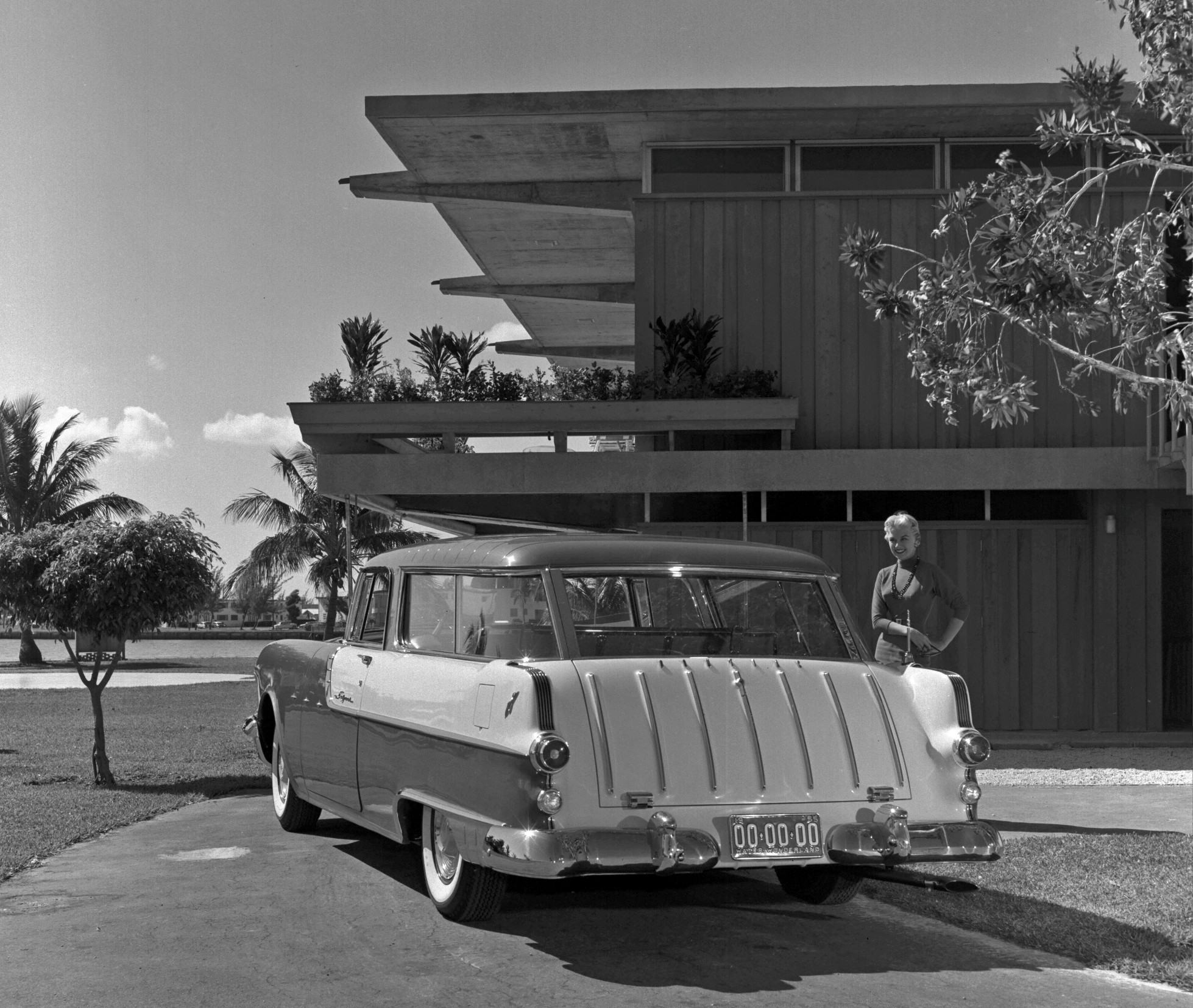 A black and white image showing a rear view of a 1955 Safari parked in front of a midcentury modern home