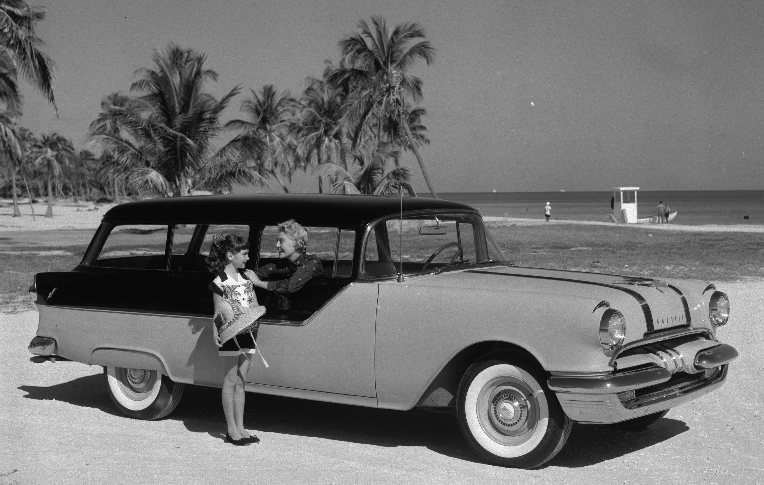 A black and white image of a two tone 1955 Safari with a black roof and with a little girl standing by the front passenger door in a beach setting with palm trees in the background and an ocean view