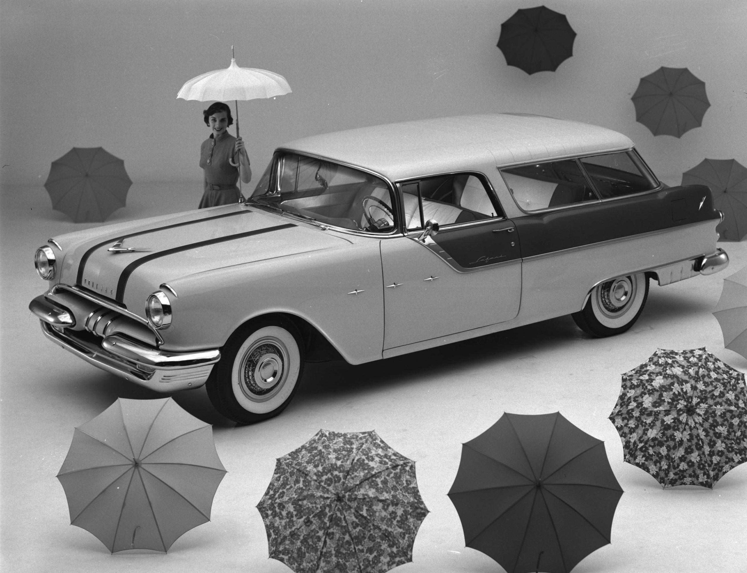 A black and white studio PR image with a 1955 Safari with a border of umbrellas and a female model holding an open white umbrella on the opposite side of the car.