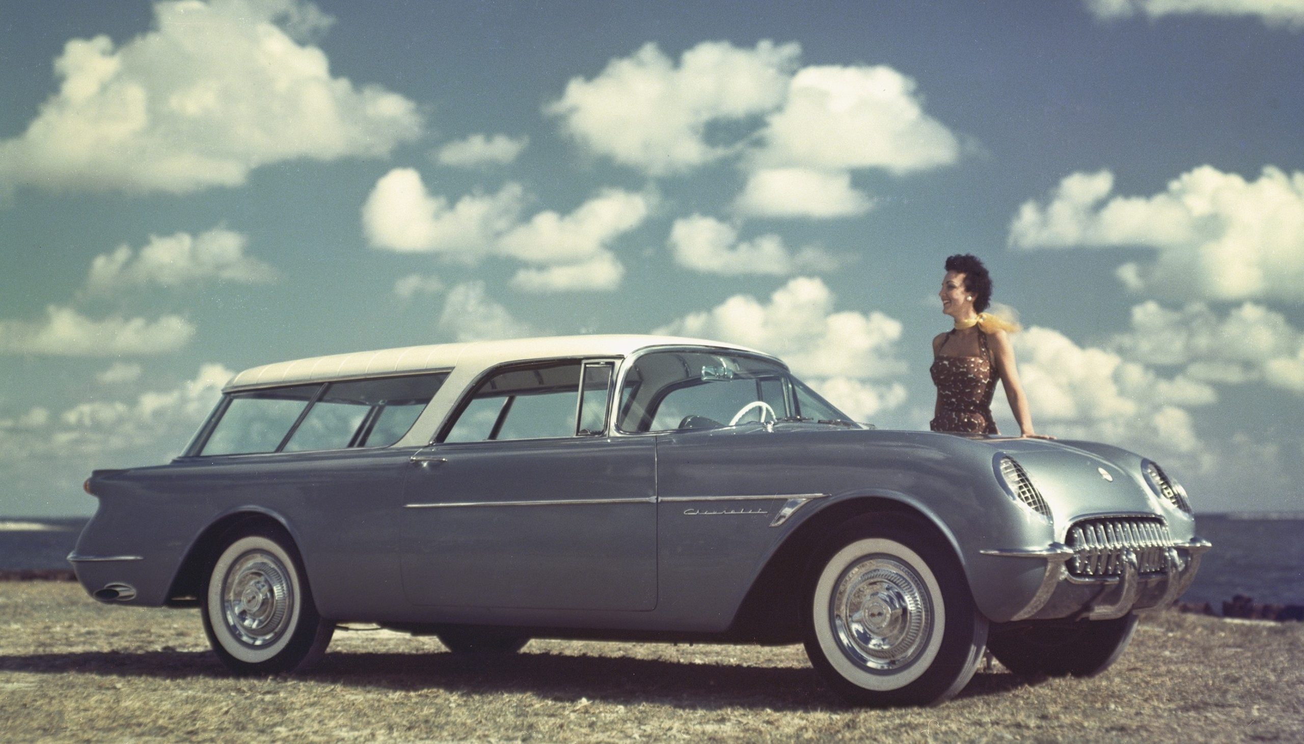 A color image of the blue 1954 Chevrolet Corvette Nomad 2 door wagon with blue skies and white clouds with a female model by the hood