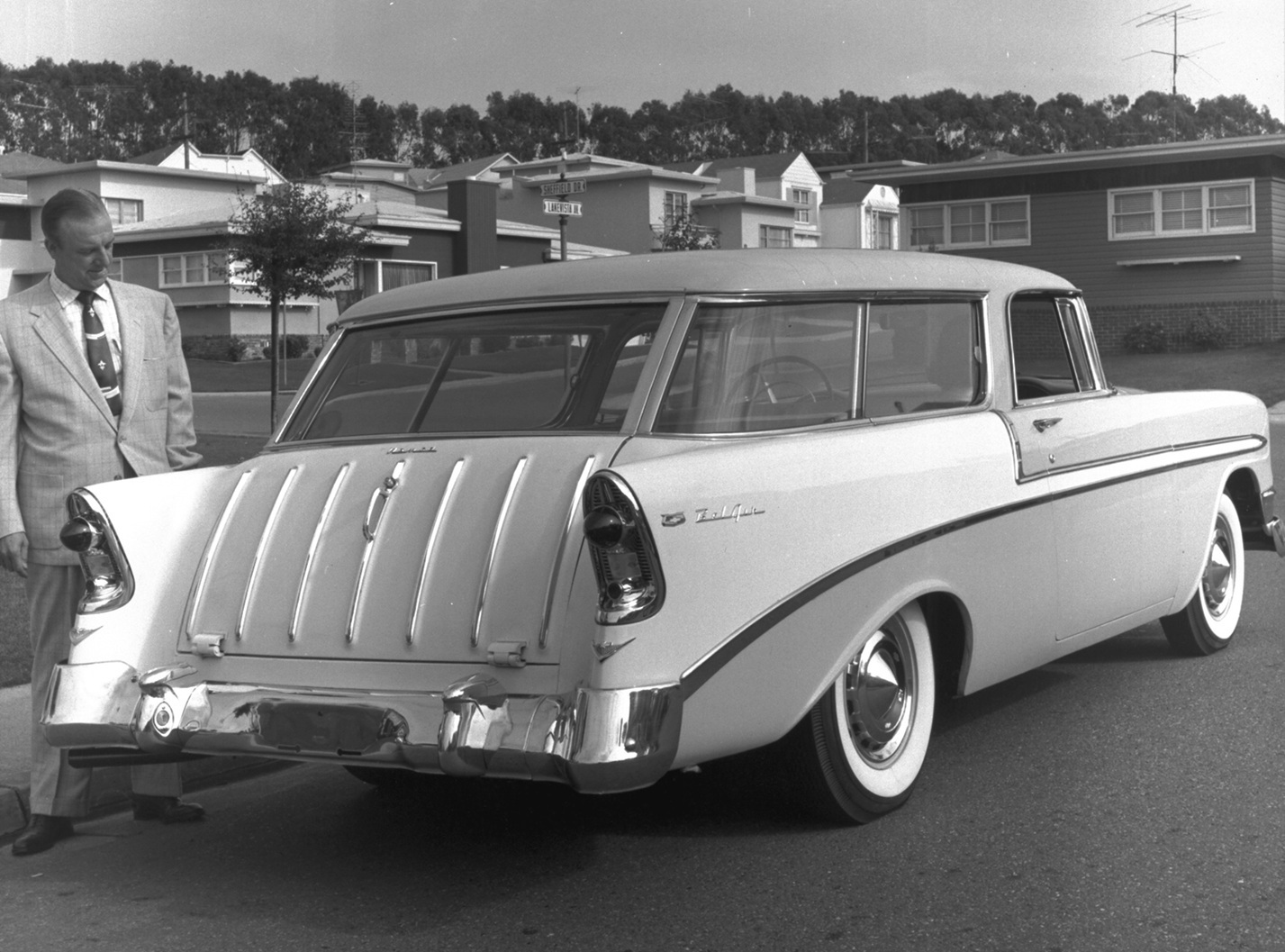 A black and image of a 1956 Chevy Nomad wagon on a suburban street showing a well dressed man admiring the car.
