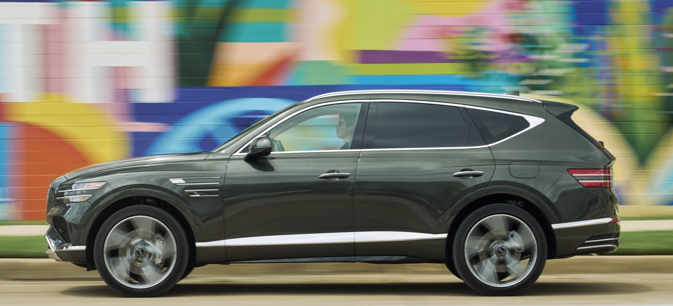 A very dark green (Storr Green) Genesis GV80 driving in front of a colorful wall