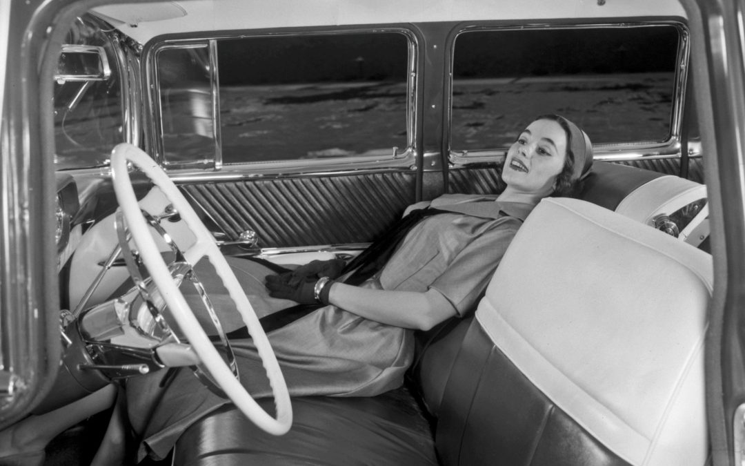 A black and white image shows a woman reclines in the front passenger seat of a 1957 Safari Transcontinental