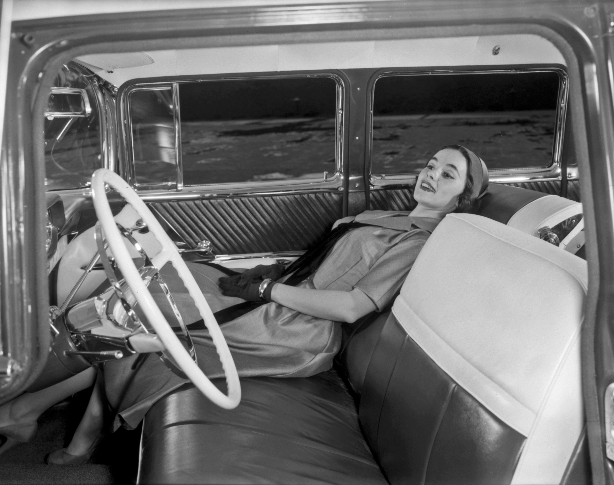 A black and white image shows a woman reclines in the front passenger seat of a Safari Transcontinental